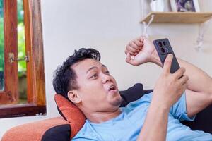 An asian man excitedly reacts while using his smartphone, relaxing on a couch near a window. photo