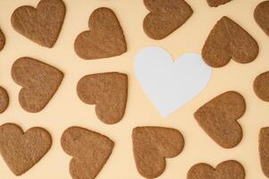 Pattern made of heart shaped cookies on cream background with paper card note copy space. Minimal love layout. Creative biscuits idea. Sweet food backdrop. Valentines day concept. Romantic flat lay. photo