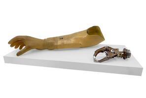 A vintage prosthetic arm with a mechanical gripper, displayed on a white pedestal. The natural-toned arm and metallic gripper highlight early prosthetic engineering and design photo