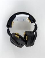 A pair of black headphones featuring gold pins integrated into the ear cushions and headband, mounted on a white wall. Designed as part of a prosthetic-related concept, blending functionality and photo