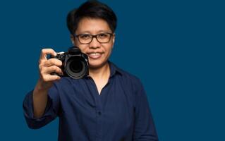 A woman photographer with short black hair holds a digital camera while standing on a blue background photo