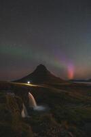 Aurora borealis dances around Kirkjufell mountain on the Snaefellsnes peninsula in West Iceland. A man stands with a lantern by Kirkjufellsfoss, watching the most unique evening show photo