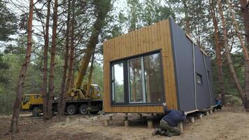 Installation of a Modern Prefabricated Cabin in a Forested Area During. The process involves precise maneuvering and placement of the structure onto a foundation amongst the tall pine trees. video