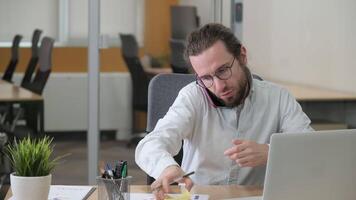 Busy man sitting on his desk at office having a talk on his cellphone, takes notes and discussing a handheld documents of marketing analytics report. Businessman doing a multi-tasking job. video