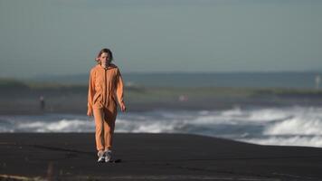 Front view of woman dressed in orange space warm jumpsuit with fleece walking on black sandy beach at golden hour. Fashion model during summer vacation. Full length, cinematic shot, real time video