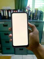 a man's hand holding a smartphone or mobile phone with a blank white screen in the office. Mockup image of smartphone with blank white screen photo