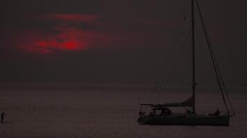Man on SUP board floating towards sailing yacht in red sunset video