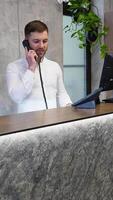 Friendly receptionist talking on phone at counter in hotel. Vertical video