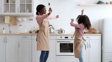 Black mother and her daughter are joyfully dancing while cooking in a modern kitchen, both wearing aprons. They are having a delightful time with utensils in their hands, showcasing family bonding. video
