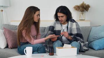 A young woman assists a man in taking medicine while sitting on a couch at home. They appear engaged in a caring moment, suggesting concern and comfort. video