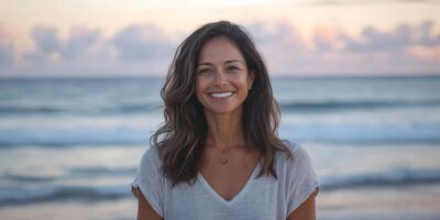 un mujer con largo marrón pelo es sonriente a el cámara en un playa. ella es vistiendo un blanco camisa y un collar. escena es contento y relajado, como el mujer es disfrutando su hora a el playa foto