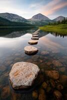 A series of rocks are in the water, with a beautiful mountain range in the background. The water is calm and still, reflecting the surrounding landscape. Concept of tranquility and serenity photo