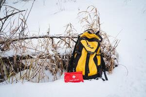 In the winter landscape, a bright yellow backpack is resting in the soft, fluffy, white snow, positioned right beside a vibrant red first aid kit, creating a beautifully striking contrast photo