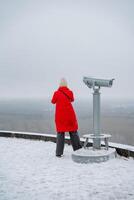 A woman is standing in the cold snow wearing a bright red coat while looking through binoculars, searching for something interesting on this chilly winter day in the snowy landscape photo