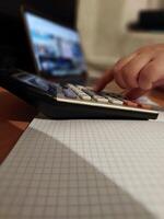 Hand using calculator on graph paper desk with open laptop in background photo