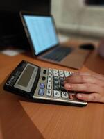 Hand using calculator at desk with laptop in background photo