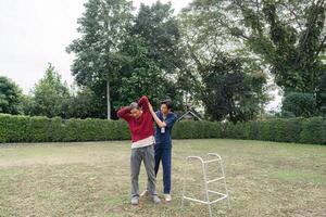 Young caregiver supporting elderly man with stretching exercise using walker in garden photo