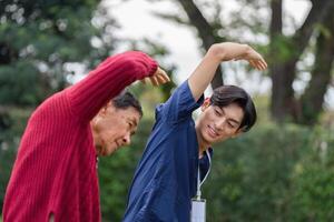 Young caregiver encouraging elderly man to perform arm stretching exercise in garden photo