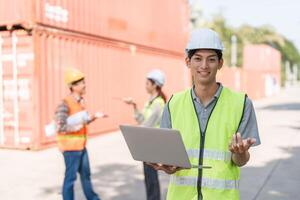Smiling construction engineer holding a laptop while coordinating with the team at a shipping container site photo