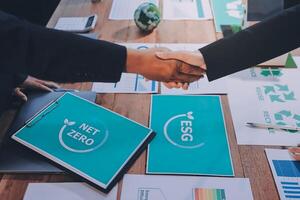 Two people shaking hands over a table with papers and a net zero logo photo