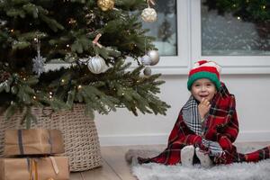 A happy little girl in a Santa Claus hat is wrapped in a warm plaid blanket by the Christmas tree photo
