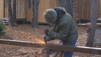A person is cutting metal using a handheld tool in a wooded setting. Sparks fly as they focus on their task under the clear sky, surrounded by trees and rustic buildings. video