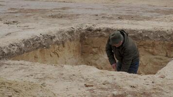 A man wearing a hat and jacket is digging a deep hole in the sandy ground. Clumps of soil are being thrown aside as he works diligently on this excavation project. video