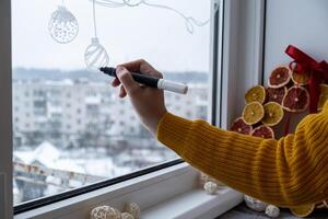 Creative enthusiastic unrecognizable woman in sweater paint window with white marker. Advent candle decoration on big clear window. Snowy winter outside. Holiday preparation photo