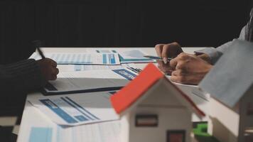 A couple of people are sitting at a table with a house model and papers video