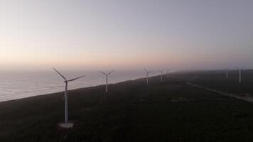 Wind turbines generate clean energy on a coastline as the sun sets and casts a warm glow on the land video