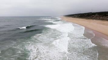 Ocean waves crash on a remote sandy beach, surrounded by vegetation and distant wind turbines under a blue sky video