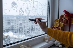 Creative enthusiastic unrecognizable woman in sweater paint window with white marker. Advent candle decoration on big clear window. Snowy winter outside. Holiday preparation photo