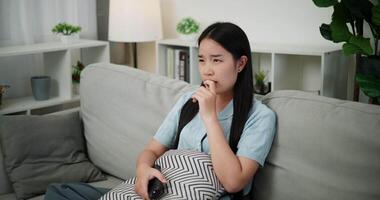 Handheld high angle view shot, Young Asian woman watching an exciting movie with a shocked and thrilled expression while sitting on a sofa in her living room at home. Leisure activity. video