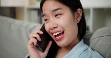 Handheld selective focus shot, Cheerful young woman is having fun talking on smartphone with her friend, smiling and laughing happily while sitting on the sofa in the living room at home. video