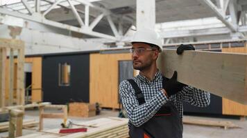 Worker carpenter in uniform walks with plank near prefabricated modular house in construction site video