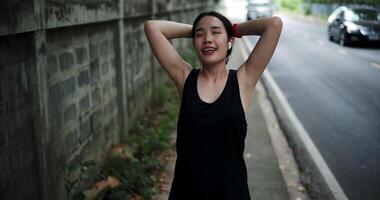 Handheld high angle view shot, Young woman in sportswear and wireless headphones listening to music while stretching to warm up before an outdoor workout on a sidewalk next to a wall in the city. video