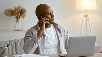 A millennial African American man in his 30s is engaged in a phone conversation while seated at his home office. He is focused, with a laptop open in front of him and papers scattered on the table. video