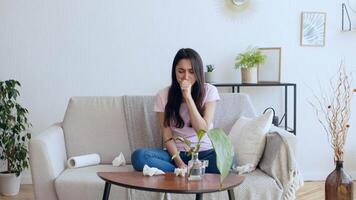 A young woman sits on a couch at home, showing signs of sickness with tissues around her. She looks unwell, reflecting the impact of flu or a cold while isolated indoors. video