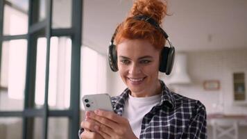 Happy smiling dancing Caucasian woman girl female at home couch weekend enjoy dance listening music in wireless headphones listen song audio sound relaxing holding smartphone looking mobile phone video