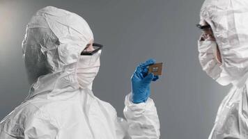 Close-up of two focused scientists in protective uniform examining, looking on new electronic chip of computer on white background. Computer CPU microchip video