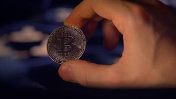 A hand is holding a shiny Bitcoin coin set against an abstract, colorful background video