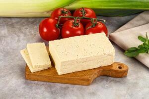 A piece of tofu on a cutting board next to a tomato and a piece of corn photo