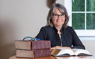 Female doctor sitting at a desk wearing glasses and a black coat with a stack of books and a stethoscope in the foreground, an open book in front of her and a window in the background. photo