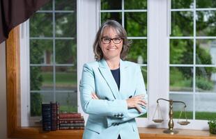 Female lawyer counselor wearing a blue coat and glasses standing in front of a window with a stack of books and a balance scale in the background. photo
