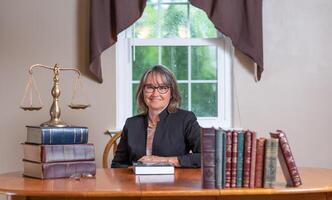 Female attorney wearing a black jacket sitting at a wooden desk between stacks of books and a balance scale with a window in the background. photo