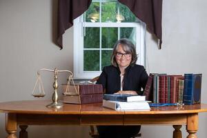 mujer abogado vistiendo lentes y un negro Saco sentado a un mesa detrás pilas de legal libros, un equilibrar escala, y un estetoscopio en frente de un ventana sonriente. foto