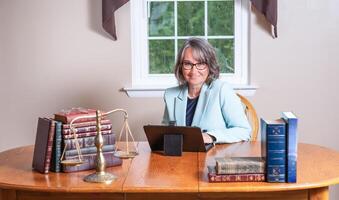 Female lawyer wearing glasses and a blue coat sitting at a laptop at a table between stacks of books and a balance scale with a window in the background. photo