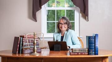 Smiling female lawyer sitting at a wooden desk with stacks of books, a balance scale, and a laptop in front of a window. photo