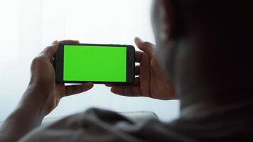 An African American man holds a smartphone with a bright green screen while sitting at home. He appears absorbed in the device, highlighting a modern digital lifestyle. video