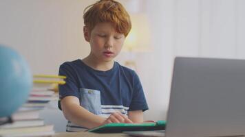 A preteen boy with ginger hair engrossed in his studies at a table. He is using a laptop and notes, focusing on his online lesson amidst a well-lit environment filled with books. video